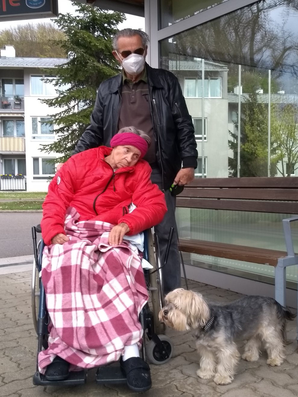 Eine Bewohnerin sitzt im Rollstuhl, neben ihr stehen ein Mann und ein Hund.